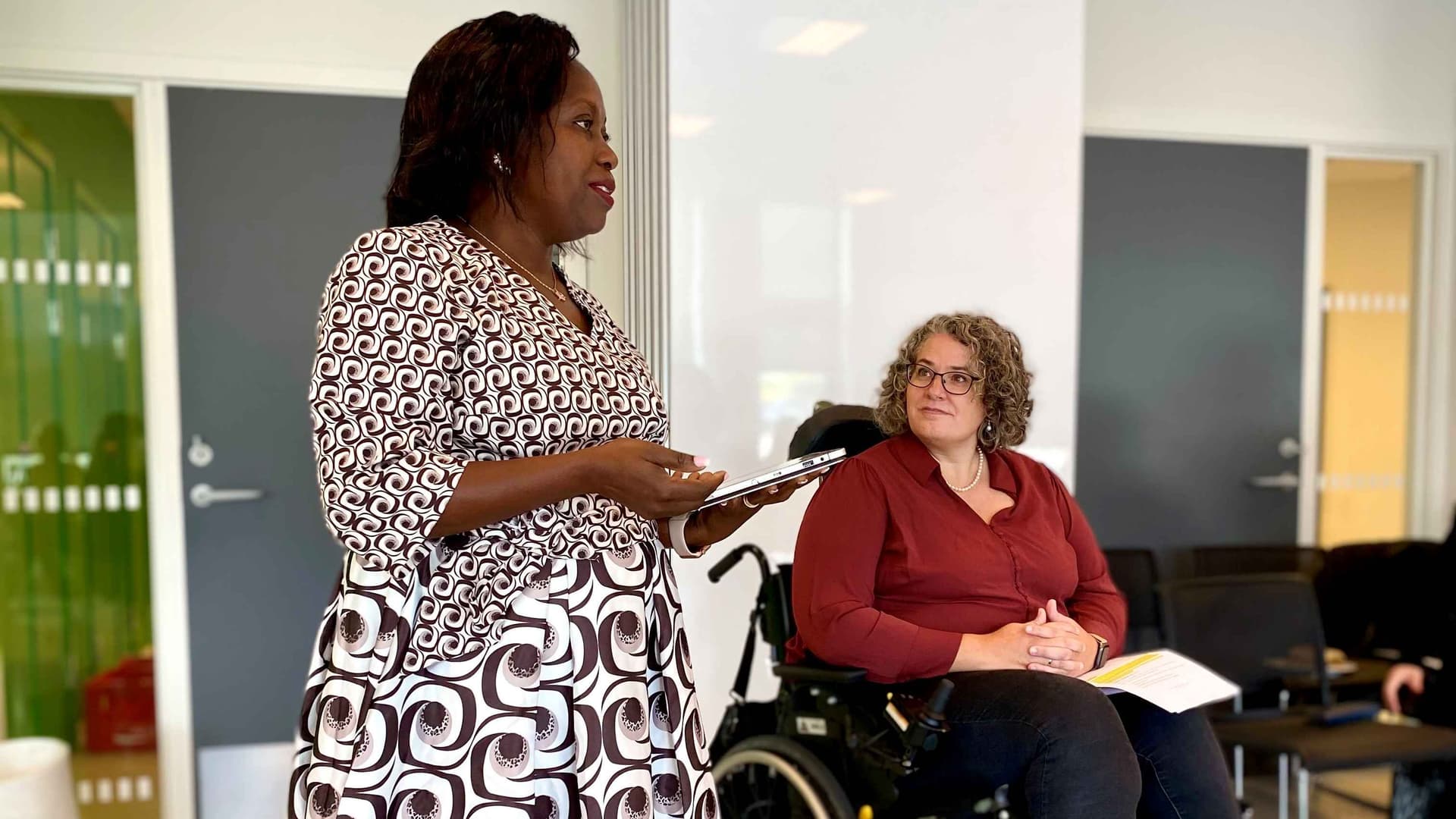 A photo of Sarah Nekesa, a beautiful black women, standing a giving a presentation. In the background, Sif Holst, a white woman in a wheelchair, is listening 