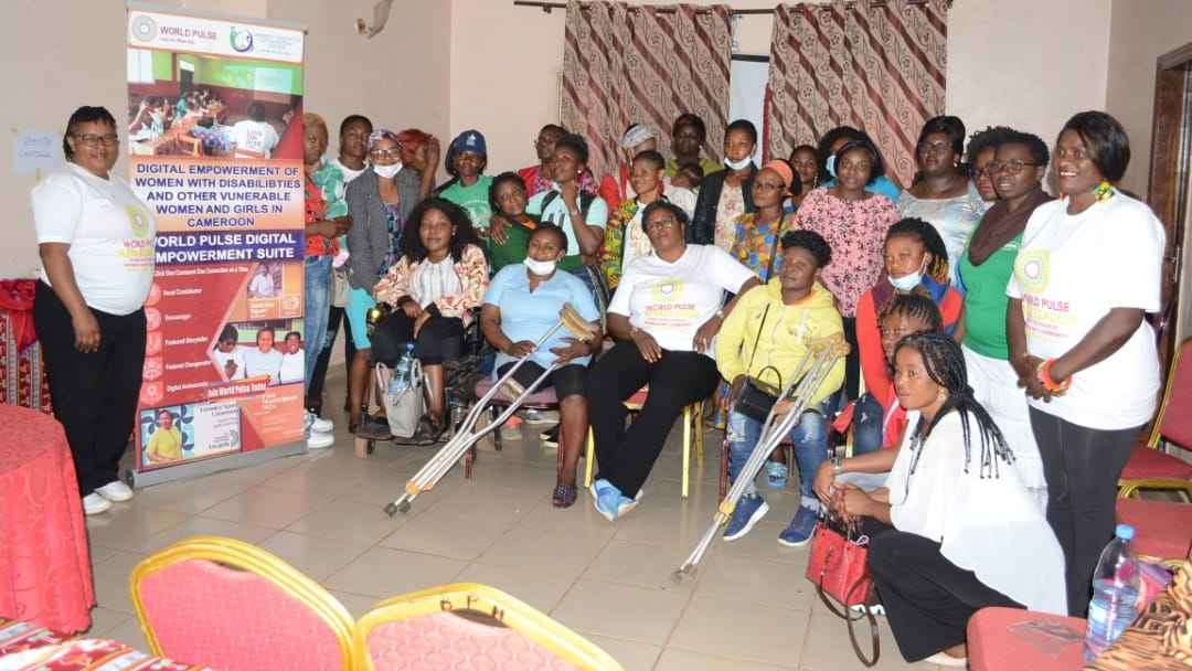 Group of women standing in a room with white tiles on the floor. Two women on the front line holding crushes and one woman sitting in a black wheelchair; A roll-up and three women wearing white World Pulse Digital Ambassador T-Shirt