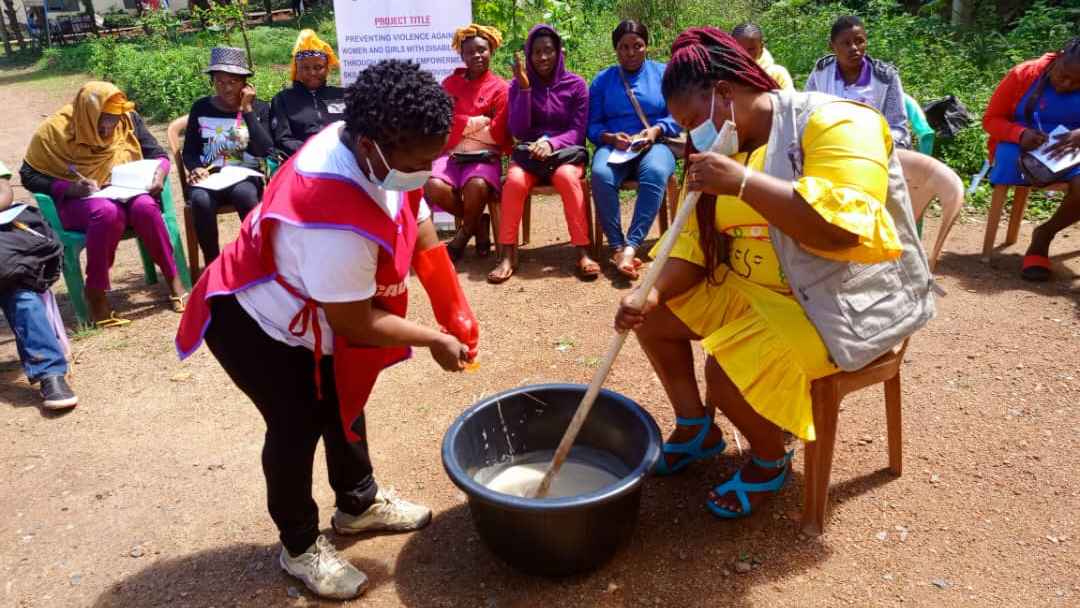 A woman, wearing a yellow dress and brown jacket, sitting on a plastic chair and stirring whitish liquid chemicals with a wooden spatula in a black basin and another woman wearing a red apron and pouring some liquid in the black basin. 