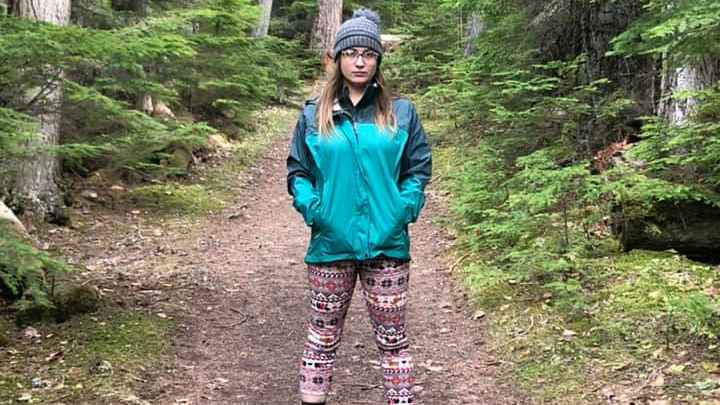 Woman with long hair, red and white pants, and teal rain jacket stands in the middle of a forest trail, surrounded by trees. She wears a floppy, grey hat on her head, and glasses. 