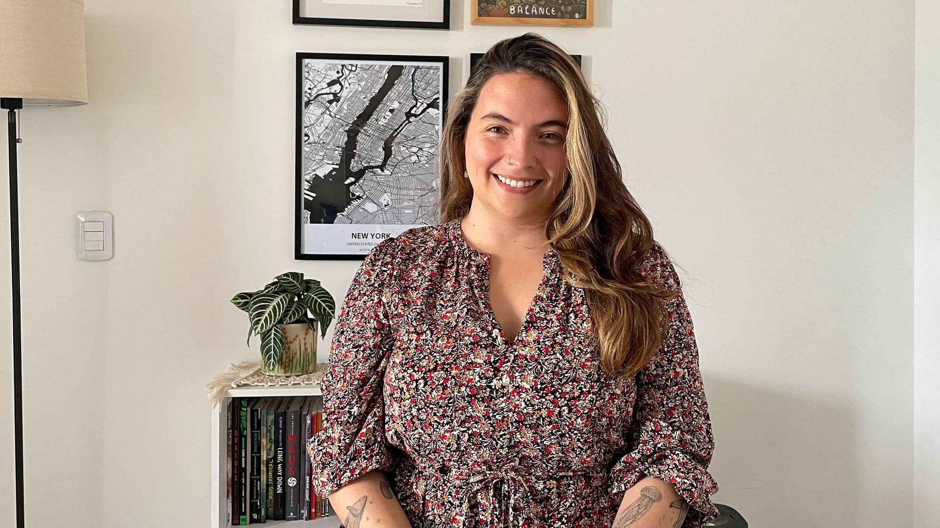 Milena Martinez, a woman with light brown hair in black, red, and cream-colored patterned shirt, is smiling in front of a bookcase with a plant and framed artwork on the wall.