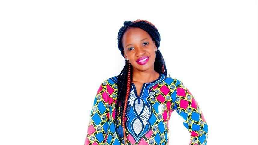 DeeRuvae has brown skin and long black braided hair with a pink streak. She smiles and has on blue eye shadow and pink lipstick. She wears a colorful blue, pink, and yellow blouse with circular shapes.