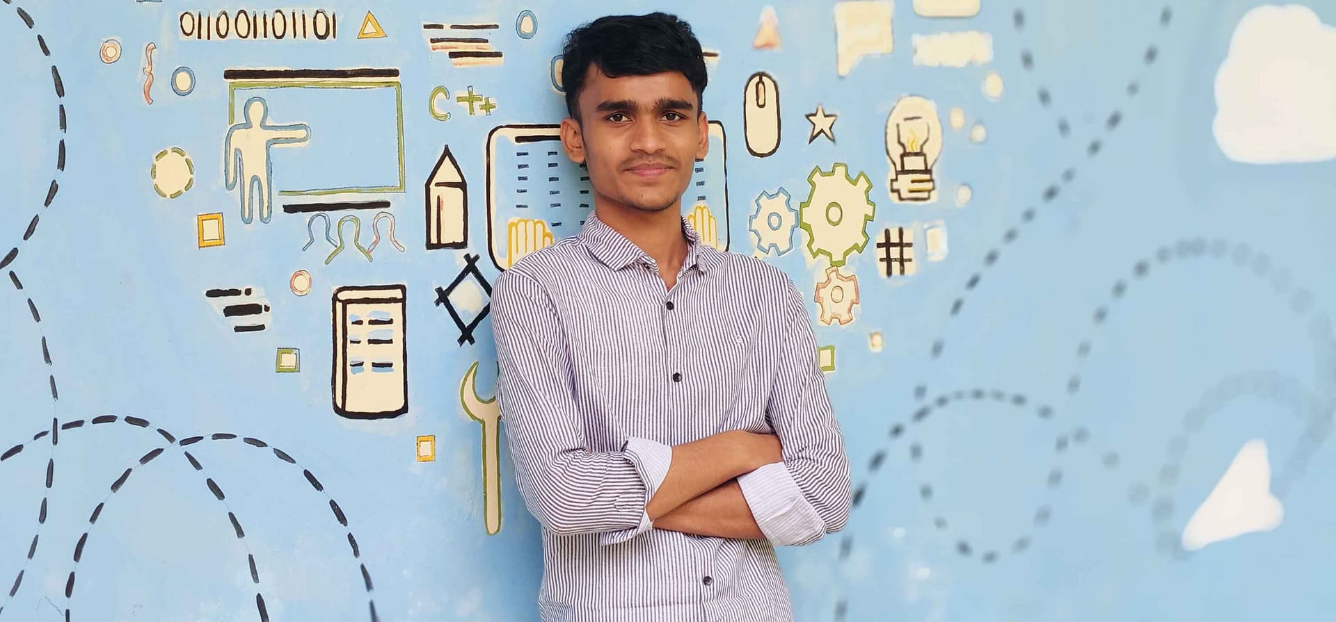 A young man standing with his arms crossed and smiling slightly, wearing a light-colored patterned shirt. He is standing in front of a blue illustrated background featuring educational and technology-related symbols.